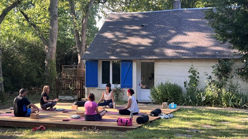 Cours de yoga en extérieur sur la terrasse en bois, devant le studio aux volets bleus