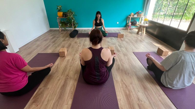 Cours de yoga collectif au studio : trois participants assis en tailleur sur leurs tapis face à la professeure